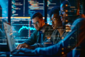 Portrait of Two Diverse Developers Using Laptop Computer Discussing Lines of Code that Appear on Big Screens Surrounding Them. Male and Female Programmers Creating Software Together and Fixing Bugs