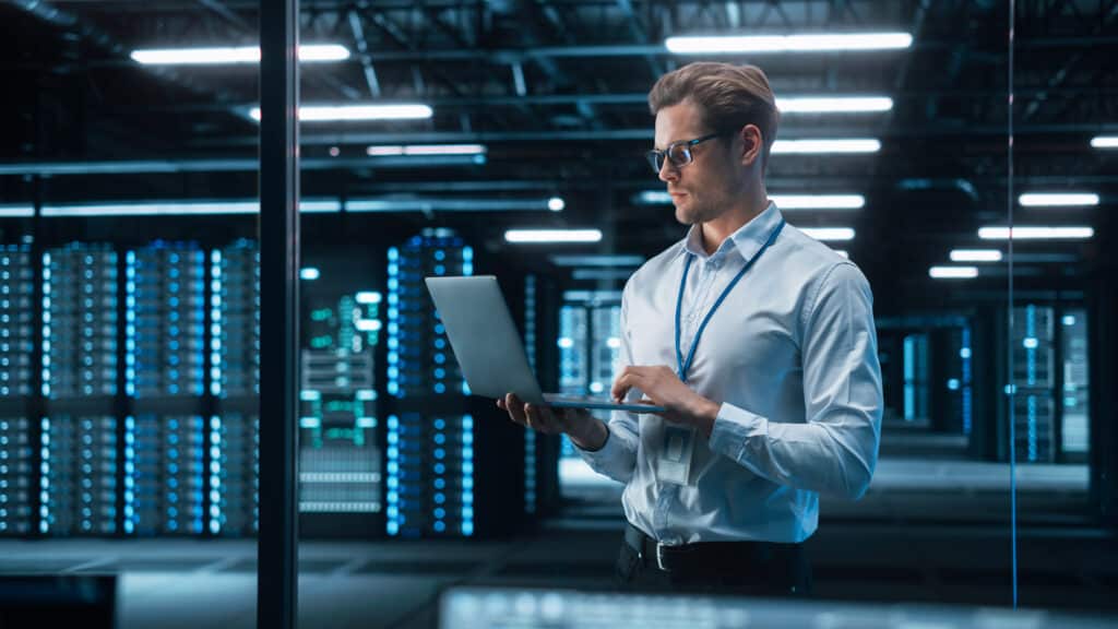 Portrait of IT Specialist Uses Laptop in Data Center. Server Far